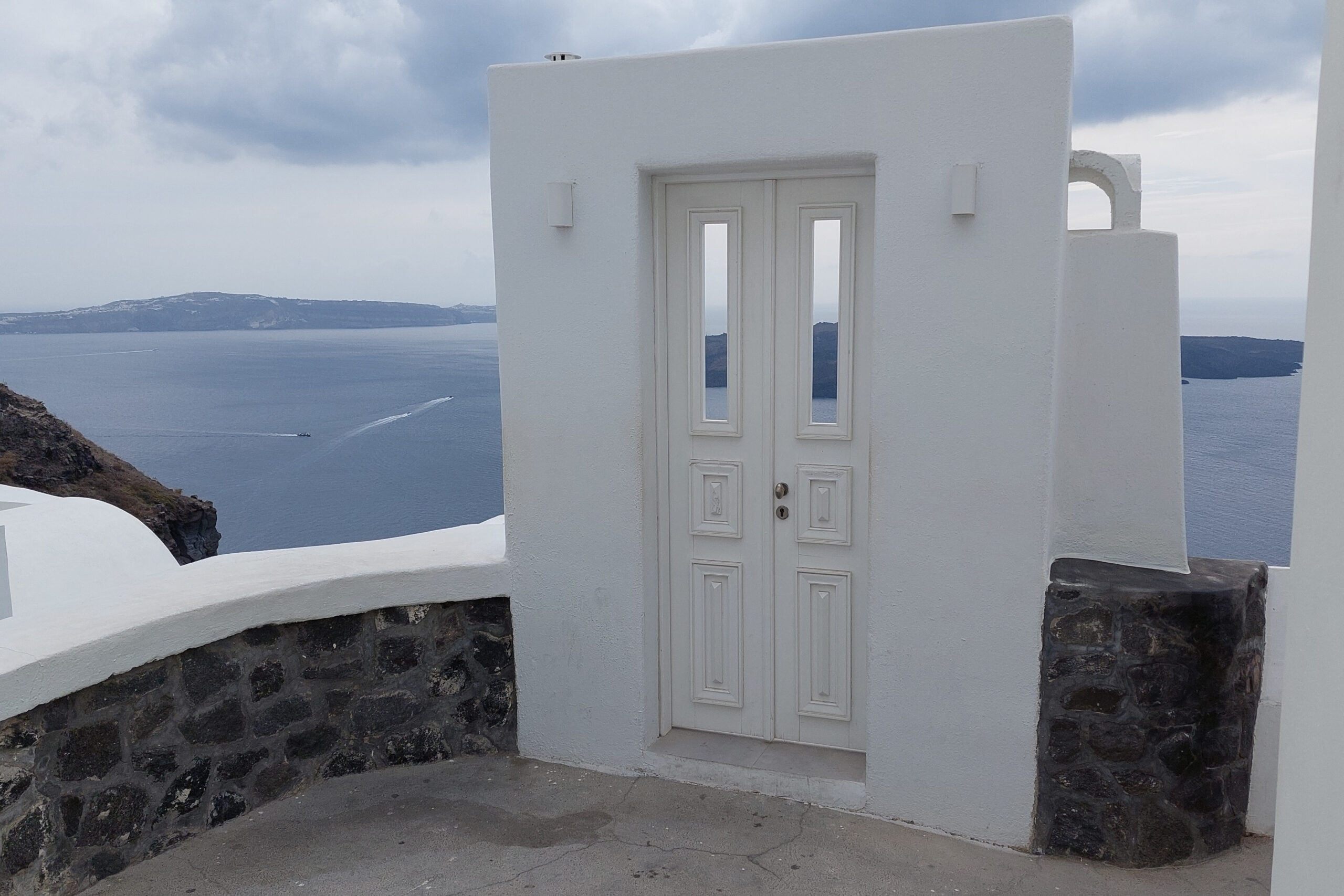 A door with two windows stands alone against dark blue water below and grey skies in the background. A short masonry wall with a white cap is visible in the foreground.