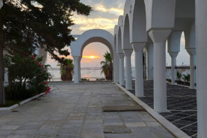 A photo of the orange morning sun peaking through scattered clouds. The sun in the middle of a white arch and more white arches are visible around the patio.