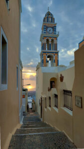 Photo looking down a cobblestone path and steps at the morning sun. A church steeple is in the center of the photo and there are dark clouds at the top of the photo.