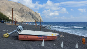 Small boat with the words "Rescue Boat" on the side in both Greek and English sits on a black sand beach. The sea is a dark navy blue. The sky is blue with scattered clouds.